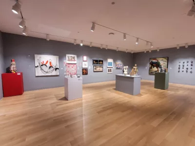 Photograph of a gallery installation with grey walls. On the perimeter walls are artworks in various mediums. There are four pedestals in the room painted various colors holding a variety of works.