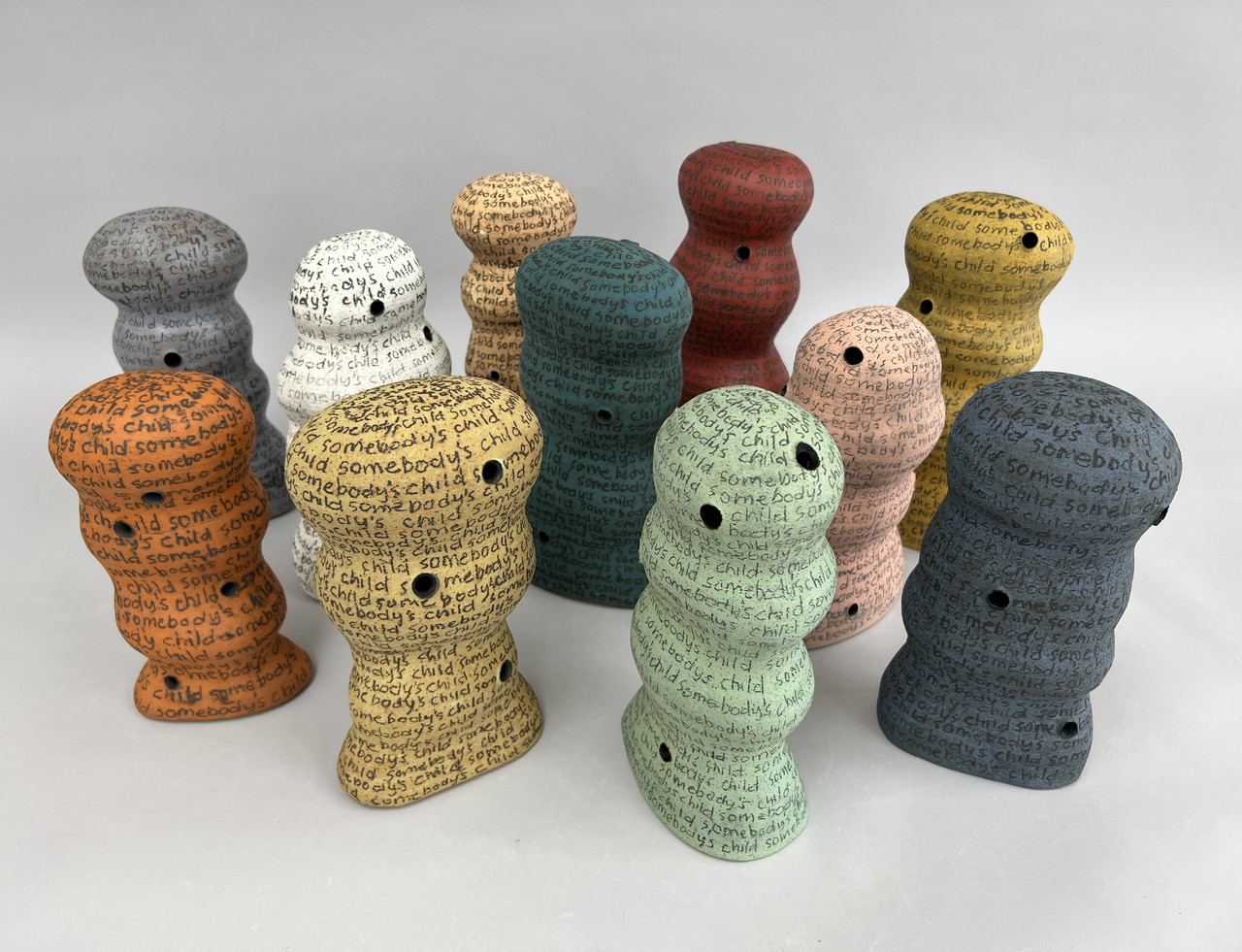 Grouping of ceramic structures assembled in a rounded ripple shape and painted in yellows, peaches, orange, greys, greens, and white colors. They have black holes and lettering around each stating 'somebody's child'.