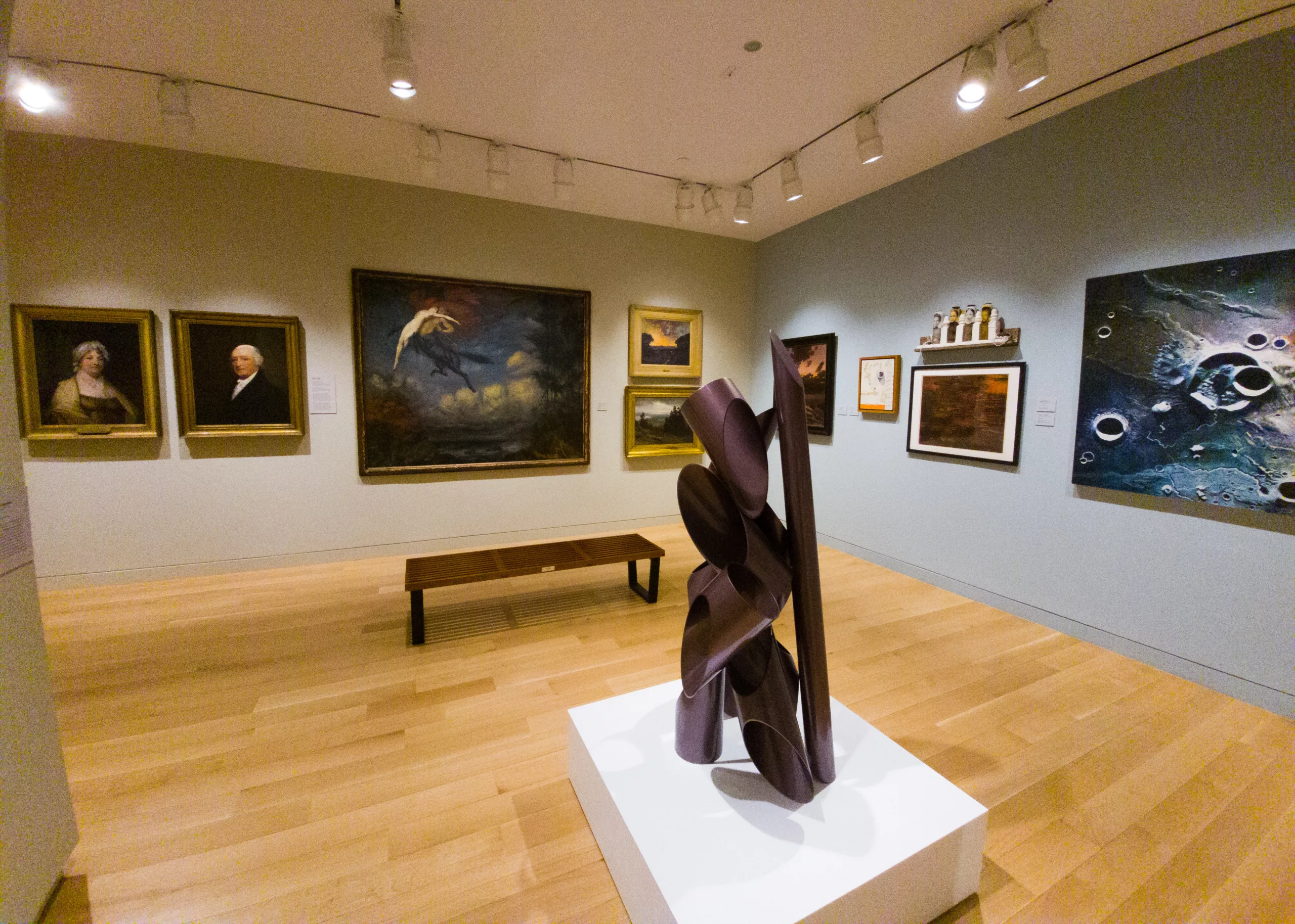 Photograph of a gallery installation with a blue wall to the right and a beige wall to the left. There are a variety of artworks hanging on the walls. In the center of the photograph is a short, white pedestal with an abstract purple sculpture made up of diagonally cut circles.