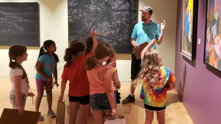 Photograph of a male teacher in a hat and bright blue shirt around a group of young children. They are standing in a gallery space around four large paintings. The girl in the back center has her hand raised.