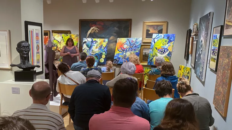 Photograph of a gallery with people seated in chairs, facing the far back wall. At the back wall is a woman standing to the left with large bright abstract paintings on easels behind her.