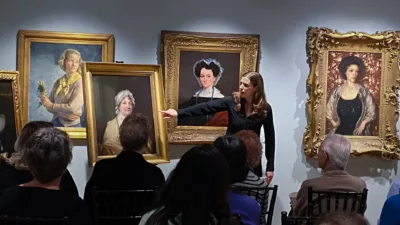 Photograph of a gallery space with many chairs and people seated for a lecture. At the front is a woman talking to the group pointing to a painting