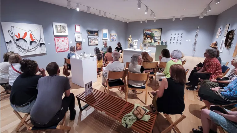 Photograph of a group of people seated in a rough circular shape in an art gallery with art on grey walls.