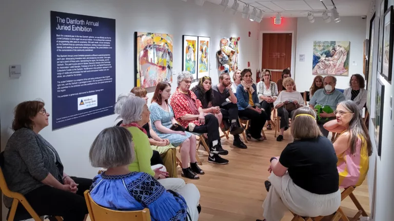 Photograph of a group of people seated in a rough circular shape in a hallway with art, facing two women to the bottom right.