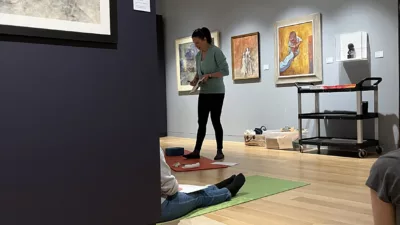 Photograph of a woman standing, talking to several women on yoga mats with paper in front of them.