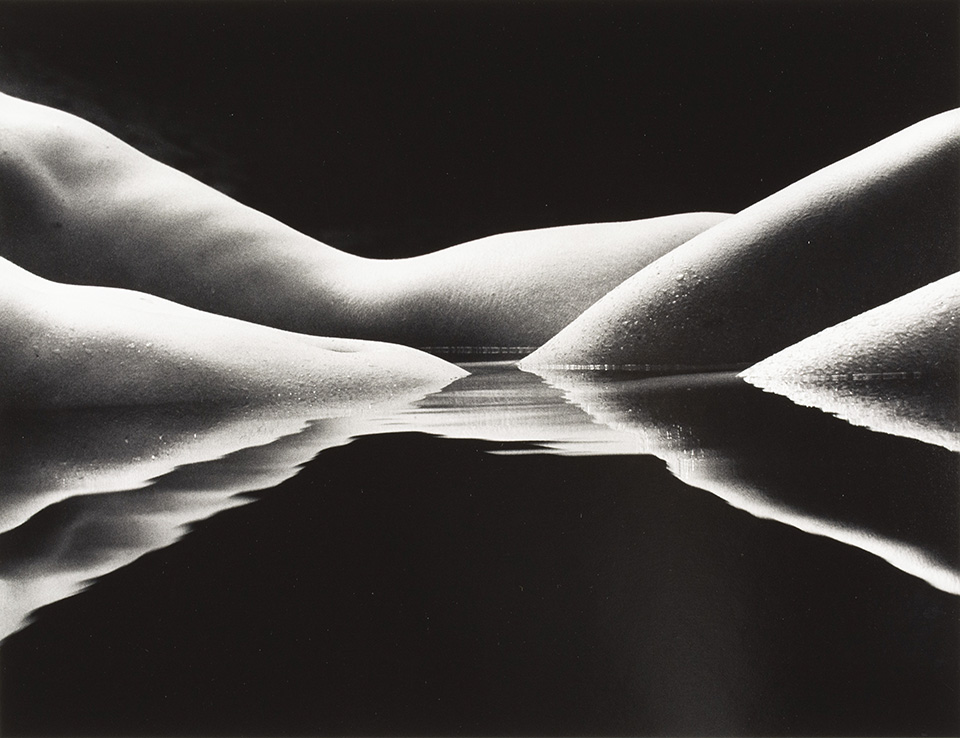 Feature image Black and white photograph of a body of water at eye level with nude human torsos and legs emerging as if they are mountain-like shapes.