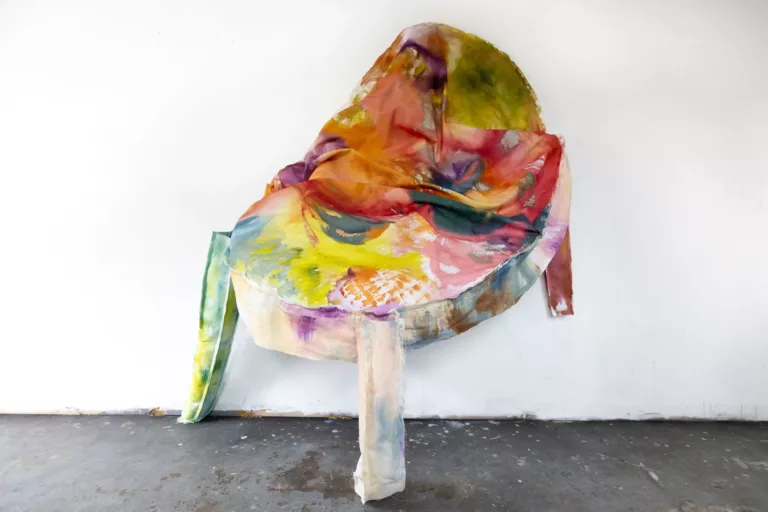 Photograph of a soft sculpture pinned to a wall and sagging downward with two arms. The fabric is brightly colored with reds, yellows, and greens.