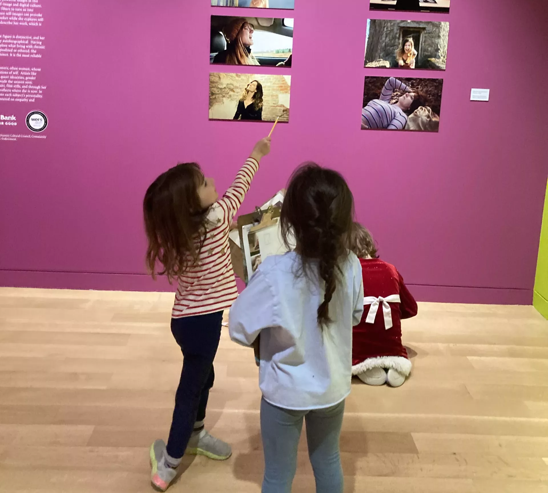 Photograph of three children with clip boards in a gallery looking at photographs.