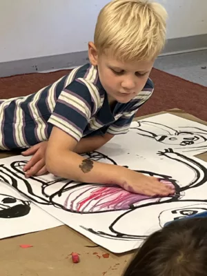 Photograph of a young boy laying on the floor drawing on white paper with black and red pastel.