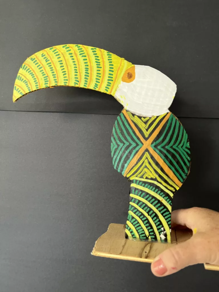 Photograph of a tucan sculpture made of cardboard and wood, painted in white, yellow, and greens.