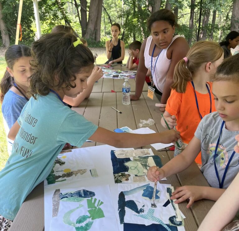 Photograph of young girls at a table, outside, painting on four sheets of paper.