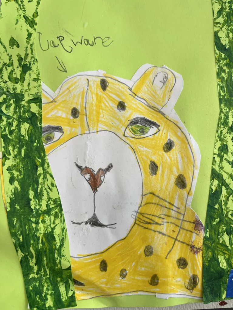 Photograph of a drawing on green paper with two strips of mottled green paper on the sides. In the center is a drawing of a spotted yellow cat's face.