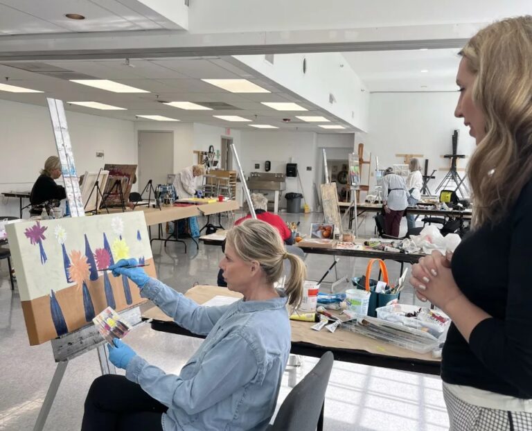 Photograph of a studio with two women in the foreground. One is seated and one standing, looking at a painting.