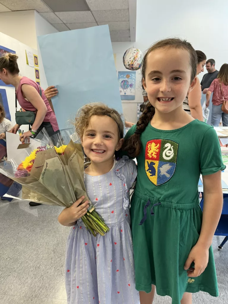 Photograph of two young girls in dresses. The girl on the left is holding flowers.