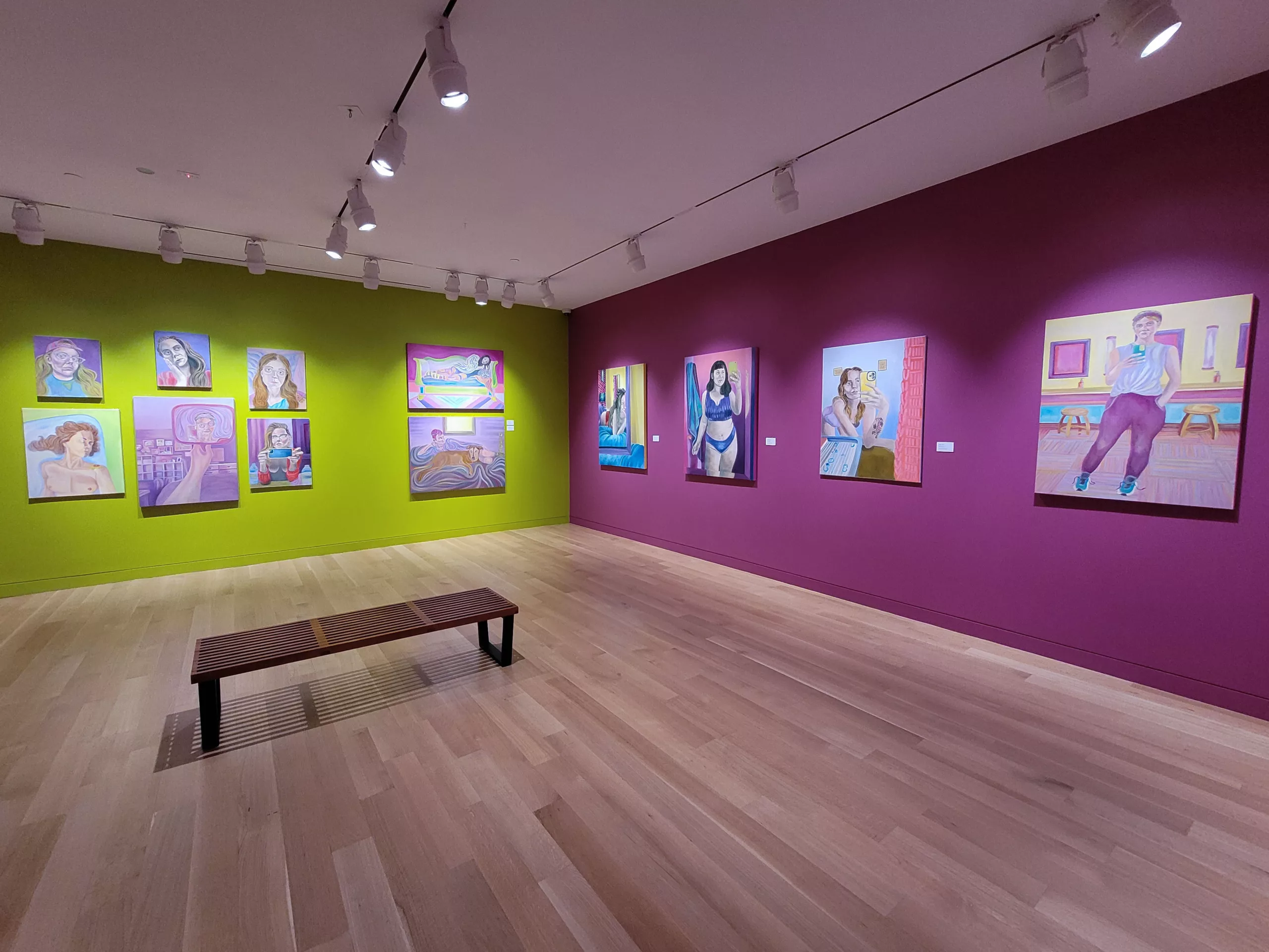 Photograph of a gallery installation with one bright green wall and one deep pink wall. Hanging are various paintings of women's portraits in bright colors.