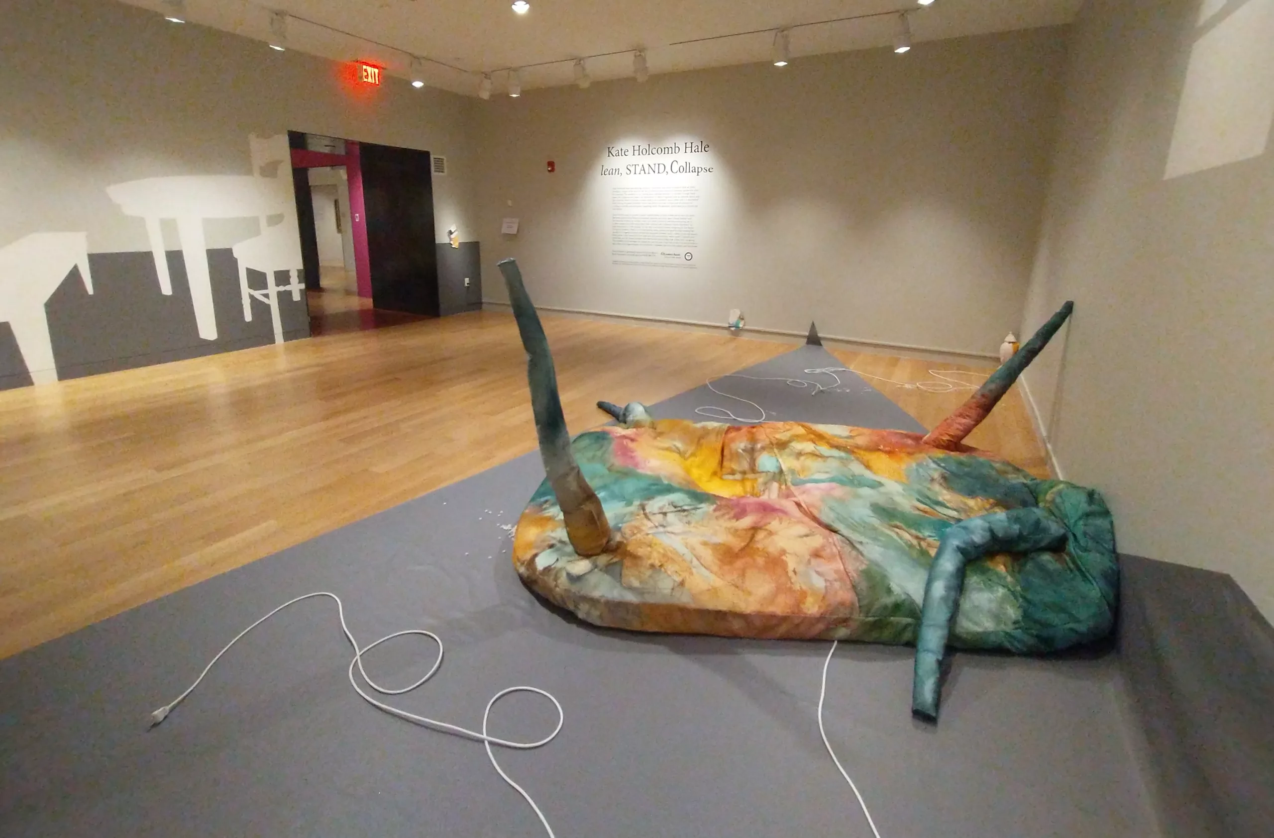 Photograph of a gallery installation with grey walls. Painted on the walls in dark grey and white is a table, chairs, and a bench. On the floor is a fabric tie dyed sculpture with three appendages on a dark grey triangular sheet.