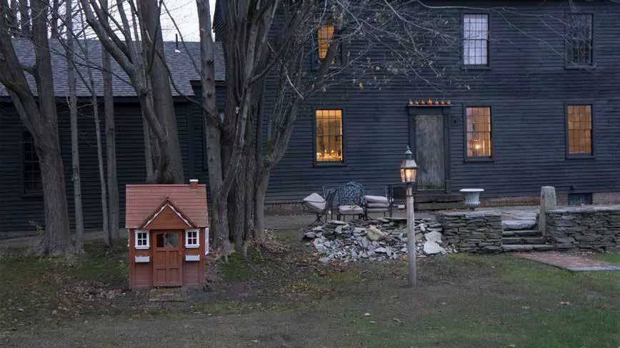 Photograph of a house with deep black siding. Several windows are lit. In front is a stone wall, a lamp on a pole, and a small red doll house to the left in front of a small grouping of trees.