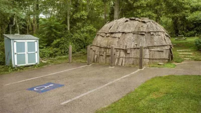 Photograph of a rounded bark house with exterior branches wrapped around. In front of this is a handicap parking spot and a small blue shed.