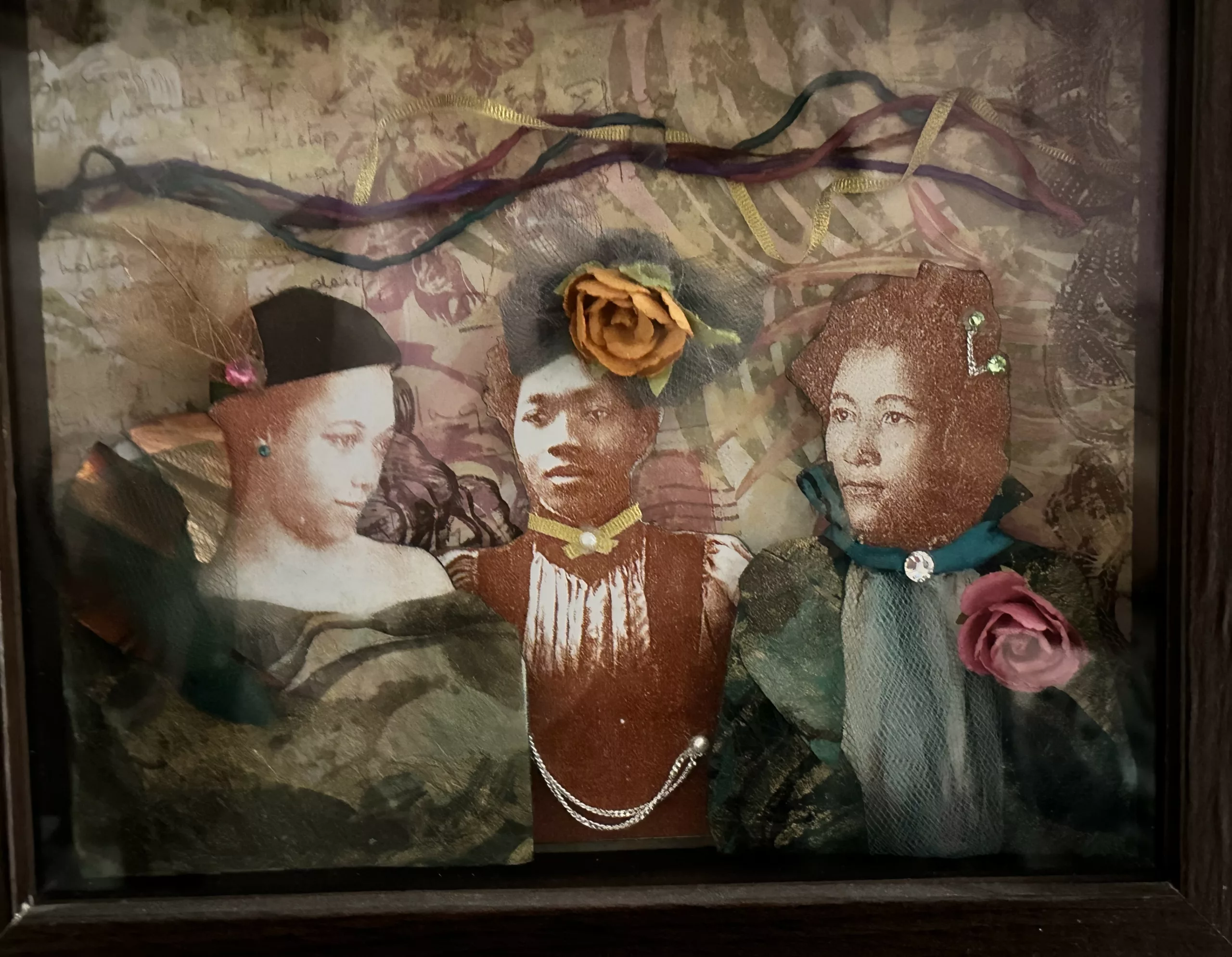 Shadow box with a leafy background in a tan color. Across the top vertically run ribbons. At the bottom are three busts of women dressed in fabric, lace, jewels, and flowers.