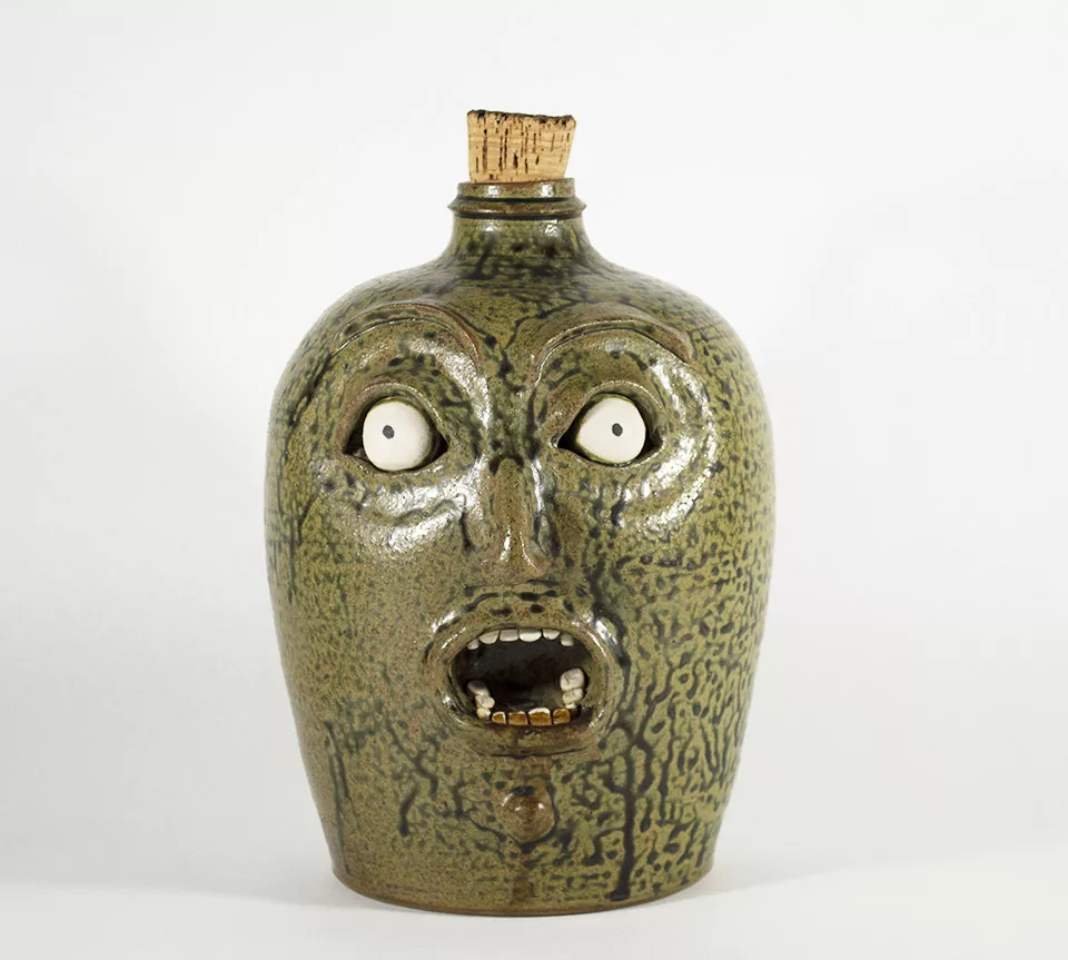 Photograph of a jug with a molted green glaze. The jug has a face with a wide mouthed expression. The top of the jug has a cork.