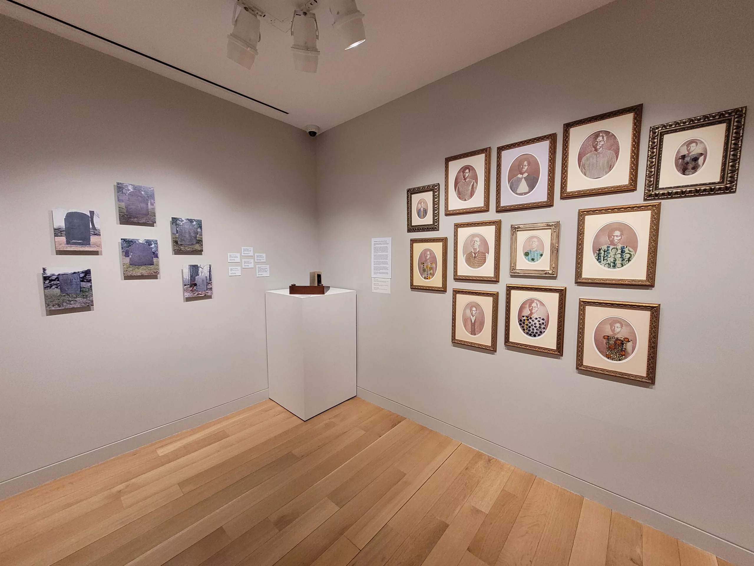 Photograph of a gallery installation with grey walls. There are photos of gravestones on the left wall, a white pedestal in between, and framed portraits on the right wall.