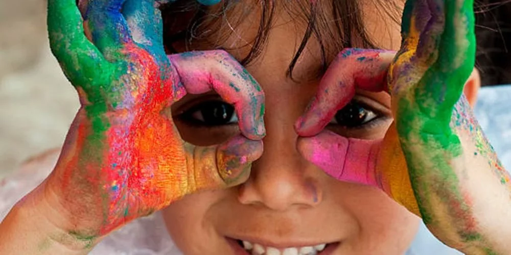 Photograph of a boy with hands up to his eyes, framing each eye. The hands are painted in bright colors.