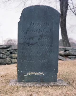 Photo of a dark grey tombstone with a broken left corner with the name Dinah at the top.
