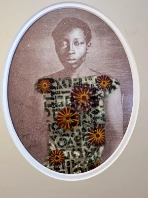 Photograph of a black and white photo in an oval mat. The subject is a woman in a floral and abstract patterned dress, colored red and green.