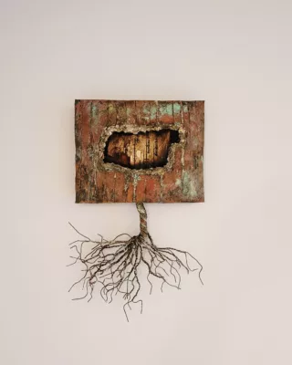 Photograph of a wall sculpture with a square-ish body in hues of red and green, and a torn section in the middle revealing tan wood. The bottom has wires coming out that look like a tree trunk and roots.