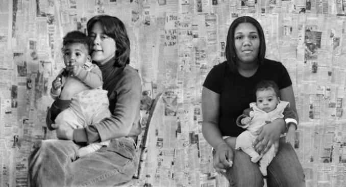 Photograph of four people seated in front of a wall of newspaper clippings. The woman on the left is holding a baby. The woman on the right is the baby, now grown up, holding her own baby.