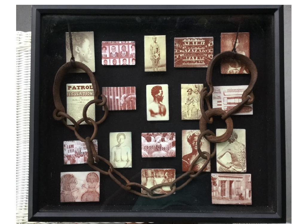 Shadow box with rectangle objects inside, each showing a photograph of an enslaved person. Across these hang a pair of slave chains.