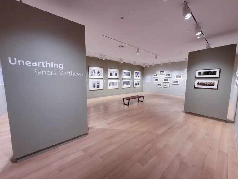 Photograph of a gallery installation with light grey walls. There are various photographs framed and hung in no particular order.