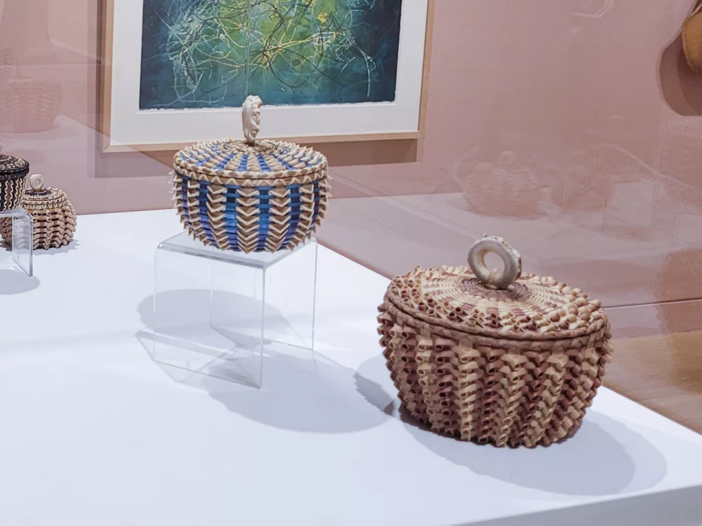 Photograph of two baskets. The one on the riser has iridescent blue stripes. The one on the right is in shades of tan and pink-tan.