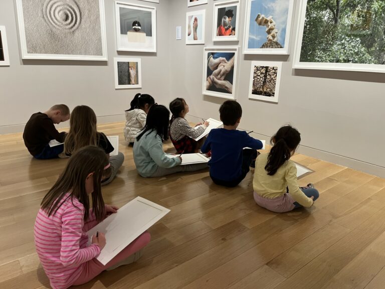Photograph of children seated in a gallery space filled with square photographs. They are drawing on large pads of paper.