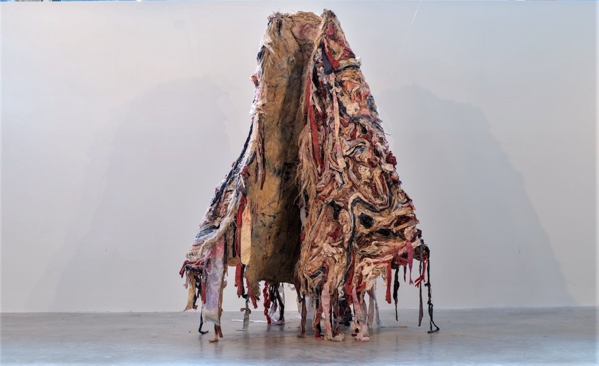Photograph of a hanging textile cloak, made with swirling red, white, black, and brown fabric.