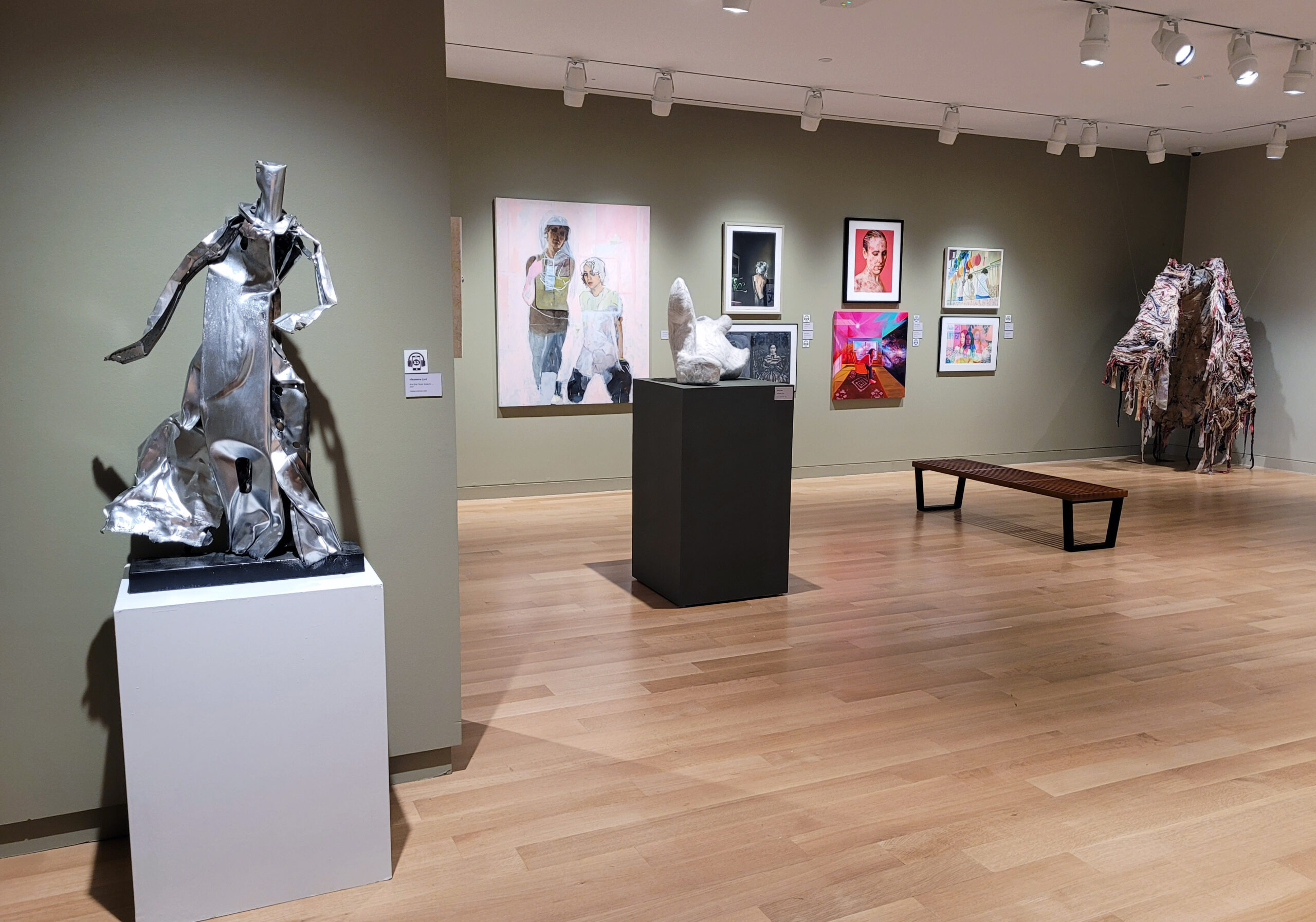 Photograph of a gallery installation with sage green walls. There is a silver metal sculpture in the front left on a white pedestal. Another pedestal and a hanging cloak piece are in the room, with artwork hanging on the wall.