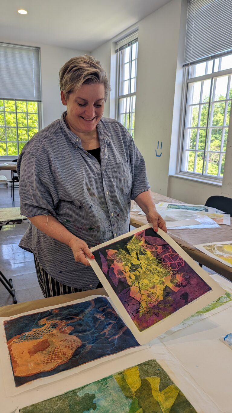 Photograph of a woman in a blue stripped collard shirt, standing behind a table, with various monotype prints in front of her.