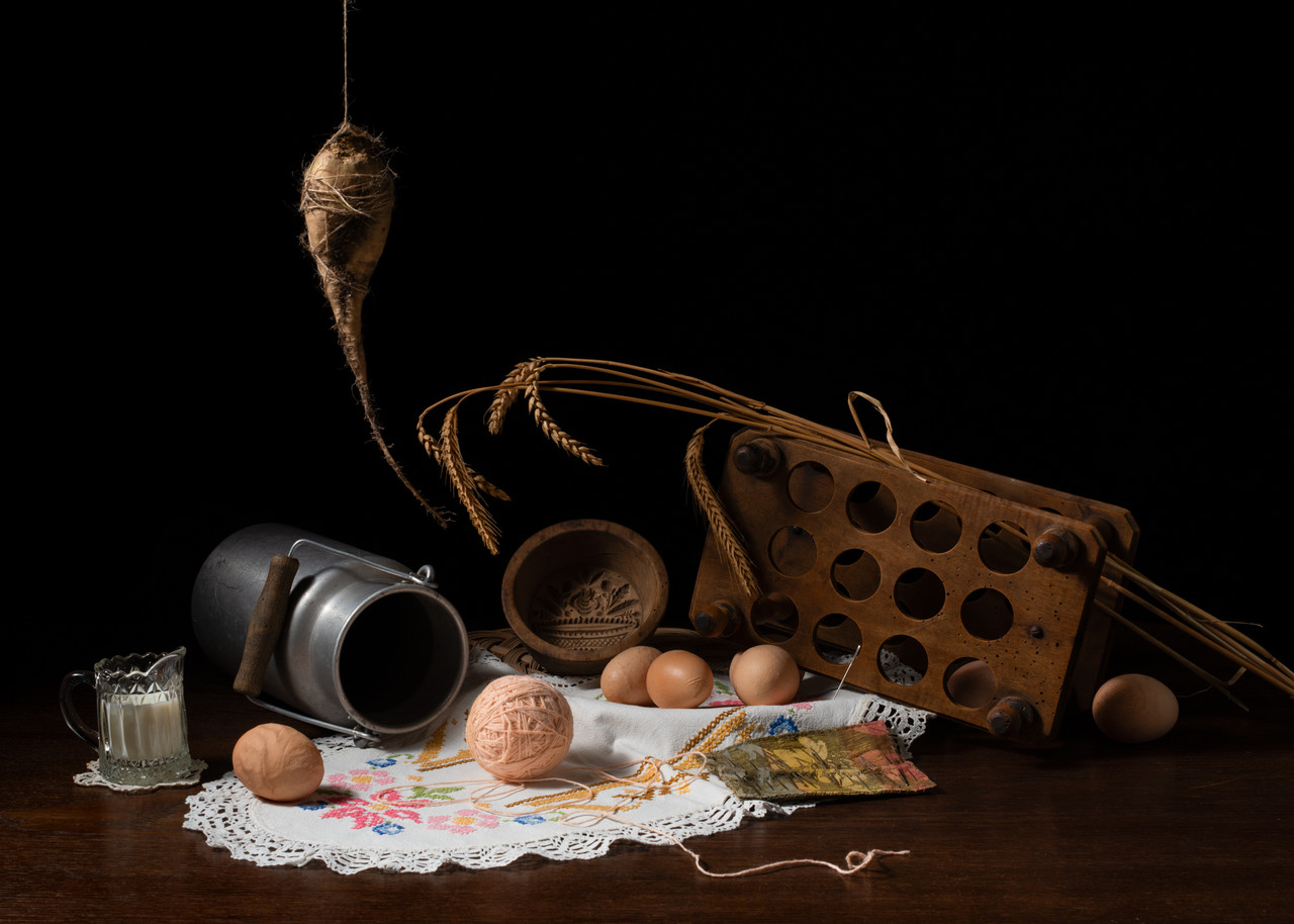 Photograph of a deep black space, with a dark brown table containing a still life of eggs, yarn, milk, milk jug, wooden objects, and wheat sheaths.