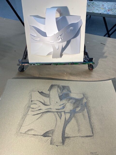 Photograph of a three dimensional paper sculpture on a small easel on a table lit with light from the left. In the foreground on the table is a drawing of the shadowed scultpure.