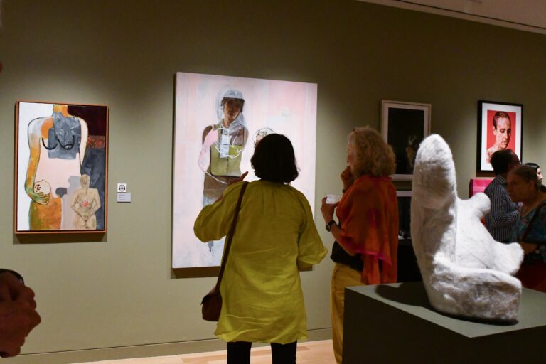 Photograph of a gallery room with sage green walls and people chatting within the room looking at the works.
