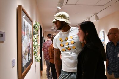 Photograph of a gallery room with cream walls and two people looking intently at one of the artworks.