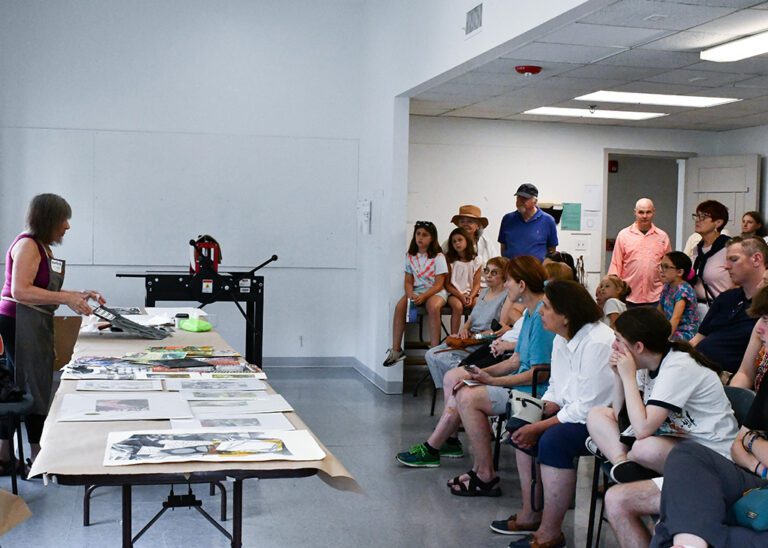 Photograph of a studio with an artist demonstrating printing on the left and a large crowd watching on the right.