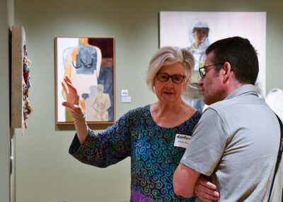 Photograph of a man and woman in a gallery talking about an artwork on the wall. The woman is gesturing to the work that is hanging on the left.