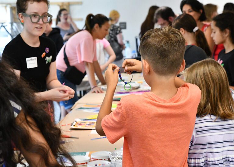 Photograph of a room filled with people gathered around a table. In the foreground is a boy facing away in an orange shirt holding up a bracelet.