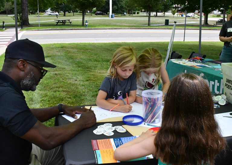 Photograph of a table outside with a man on the left and three girls on the right drawing on paper.