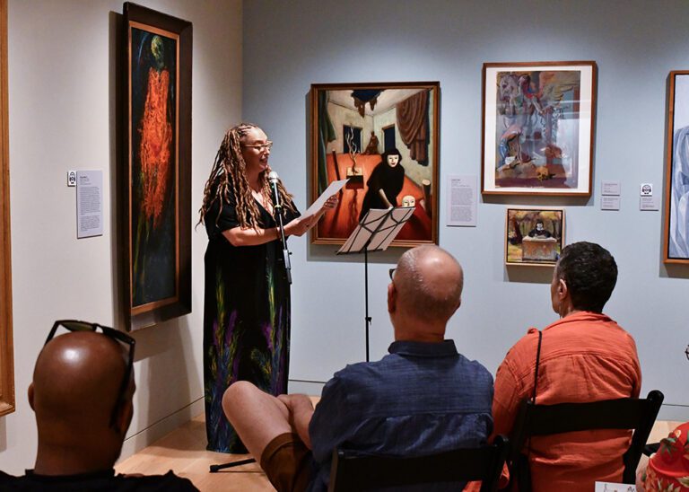 Photograph of a gallery room, with a woman standing at a mic in the back corner reading from a paper. There are people seated around listening to her read.