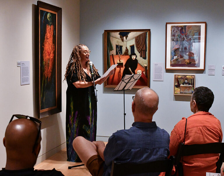 Photograph of a gallery room, with a woman standing at a mic in the back corner reading from a paper. There are people seated around listening to her read.