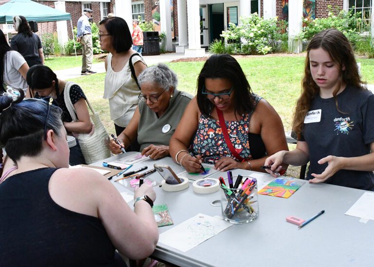 Photograph of a group of people seated at tables outside, drawing on plexiglass.