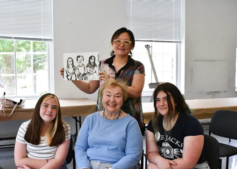 Photograph of a studio, with three women seated in front, and an artist standing behind holding up a drawing of those three women.