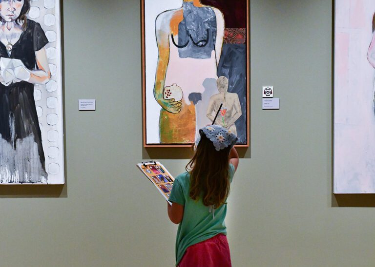 Photograph of a gallery room with sage green walls and three paintings hanging. In the foreground is a young girl with a pencil and a scavenger hunt looking at the paintings.